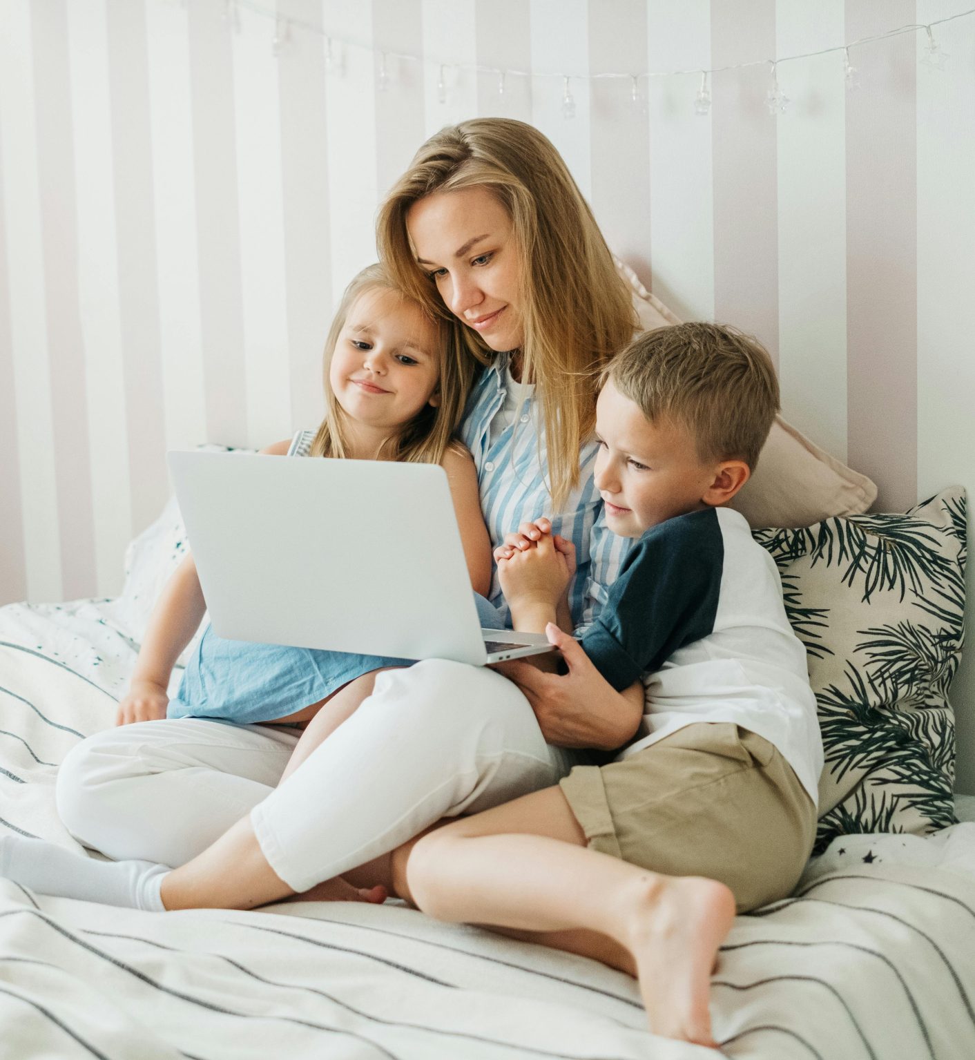 A mother sits on a bed with her children, sharing a joyful moment using a laptop.